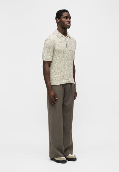 Άντρας που στέκεται, φοράει ένα μπεζ πλεκτό πόλο με κοντά μανίκια και υφή, άνετο καφέ παντελόνι και δίχρωμα μπεζ και μαύρα παπούτσια.