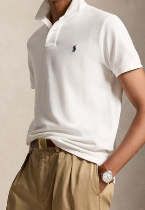 Persoon die een wit poloshirt met een klein logo draagt, beige broek met een riem en een horloge met een bruine leren band.