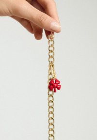 Kullanımda büyük halkalara sahip parlak kırmızı çiçek şeklinde bir charm ile bağlı altın metal zincir. Charm, karmaşık detaylara sahip.