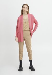 Cardigan oversized rosa con tasche, indossato sopra un top beige con pizzo e pantaloni beige, abbinato a stivali alla caviglia neri con lacci.