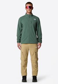 Miestna zelená, zipsová softshellová bunda s vysokým golierom. Béžové cargo nohavice so zipsovými vreckami. Čierne turistické topánky. Slnečné okuliare s červeným nádychom.