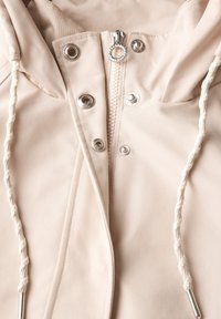 Beige huva jacka med dragkedja, silverdetaljer, öglor och dragsnören. Har strukturerad tyg och synliga sömnadsdetaljer.