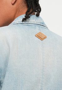 Ανοιχτό μπλε τζιν μπουφάν με υφή, το οποίο διαθέτει μία καφέ δερμάτινη ετικέτα με το λογότυπο "REPLAY" στο πίσω κολάρο.