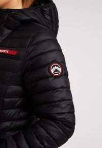 Giacca a vento nera con cappuccio, cuciture orizzontali, dettagli in logo rosso e un patch circolare con grafiche montane sulla manica.