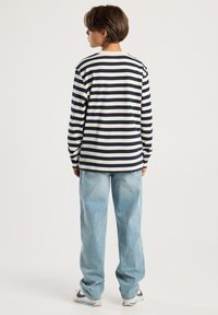 Kind, der mit dem Rücken zur Kamera steht, trägt ein langärmliges schwarz-weiß gestreiftes Shirt, hellblaue Jeans und weiße Sneakers mit schwarzen Swooshes.