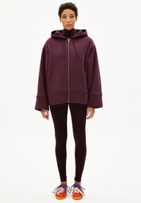 Felpa con cappuccio viola oversize con zip e tasche anteriori, abbinata a leggings a righe zebra nere e viola e sneakers multicolori.