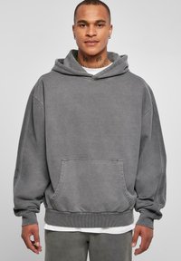 Felpa oversize grigia con tasca a marsupio frontale, cappuccio con cordino, polsini a coste e morbido tessuto di cotone. Indossata sopra una camicia bianca.