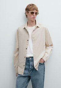 Beige kordjacka med knäppning framtill, bärs över en vit t-shirt. Modellen har på sig ljusblå denimjeans och grönt tonade solglasögon.