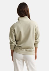 Γυναίκα με μακριά καστανά μαλλιά, ντυμένη με μπεζ πουλόβερ με γυρισμένα μανίκια και άσπρο παντελόνι, βλέποντας από την πίσω πλευρά σε απλό φόντο.