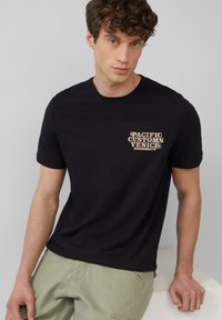 Zwarte katoenen T-shirt met korte mouwen, ronde hals, met een voorop gedrukt ontwerp dat "PACIFIC CUSTOMS VENICE BOARDS&KITS" in lichtbruin weergeeft.