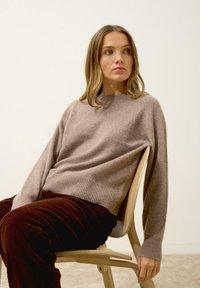 Jasnobrązowy, dziany sweter z rybym brzegiem i mankietami, w zestawie z ciemnobrązowymi spodniami z kordurowej tkaniny. Model siedzi na drewnianym krześle.