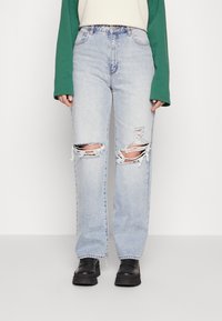 Jeans de ganga azul claro com grandes rasgões nos joelhos, corte reto, cintura alta e tecido desgastado. Combinados com sapatos pretos de sola grossa.