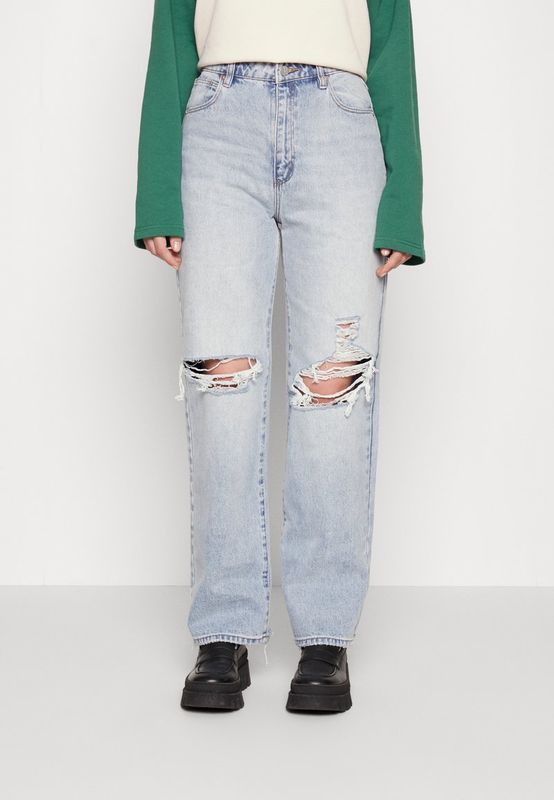 Jeans de ganga azul claro com grandes rasgões nos joelhos, corte reto, cintura alta e tecido desgastado. Combinados com sapatos pretos de sola grossa.