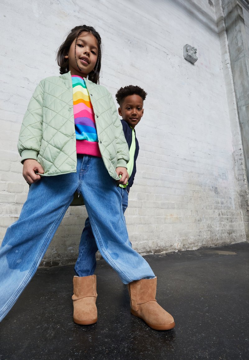 Deux enfants posent ensemble. La fille porte une veste bomber matelassée vert clair, un pull rayé coloré et un pantalon en jeans large bleu ; elle a des bottes marron. Le garçon porte une veste sombre avec des accents néon et un jean.