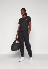 Camiseta preta e calças de treino com riscas brancas laterais, a textura do tecido é suave. Inclui uma mochila de ginástica preta. Ténis brancos completam o visual.