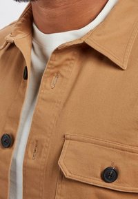 Koszula na guziki w kolorze jasnobrązowym wykonana z teksturowanej tkaniny, z ostro zakończonym kołnierzykiem, czarnymi guzikami oraz kieszenią na piersi z klapką.