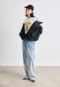 Μαύρο φουσκωτό μπουφάν πάνω από λευκή μπλούζα με κίτρινο σχέδιο, σε συνδυασμό με ανοιχτό μπλε φαρδιά τζιν και σκούρα μπλε μυτερά παπούτσια. Μαύρο καπέλο.