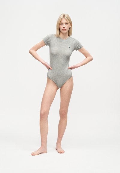 Ξανθιά γυναίκα, όρθια ξυπόλητη με τα χέρια στους γοφούς, φοράει εφαρμοστό γκρι ολόσωμο κορμάκι με κοντά μανίκια, σε απλό λευκό φόντο.