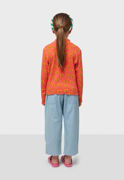 Jersey naranja con patrón de flores amarillas, pantalones sueltos azul claro y sandalias rosas. Cabello en una coleta con calentadores de orejas a rayas.