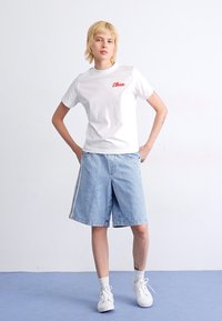Hvit bomull T-skjorte med rødt logo, parret med lyseblå denimshorts. Personen står selvsikkert, iført hvite sneakers og anklestrømper.