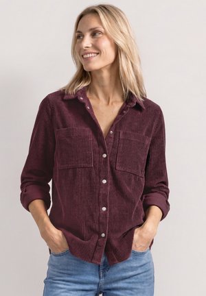 Kvinde iført en bordeaux fløjlsbluse med opkørte ærmer og blå jeans, der smiler og kigger til siden med hænderne i lommerne.