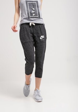 Osoba ubrana w szary top bez rękawów i czarne krótkie spodnie dresowe Nike oraz białe sportowe buty, idąca do przodu na jednolitym tle.