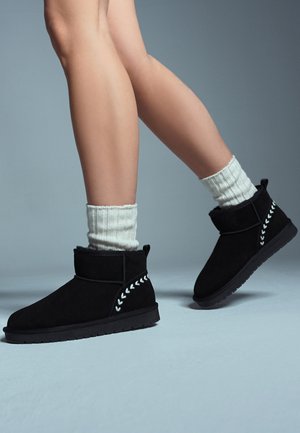 Stivaletti neri in suede con una texture morbida, caratterizzati da dettagli cuciti bianchi ai lati e una suola in gomma resistente. Indossati con una calza color crema a coste.