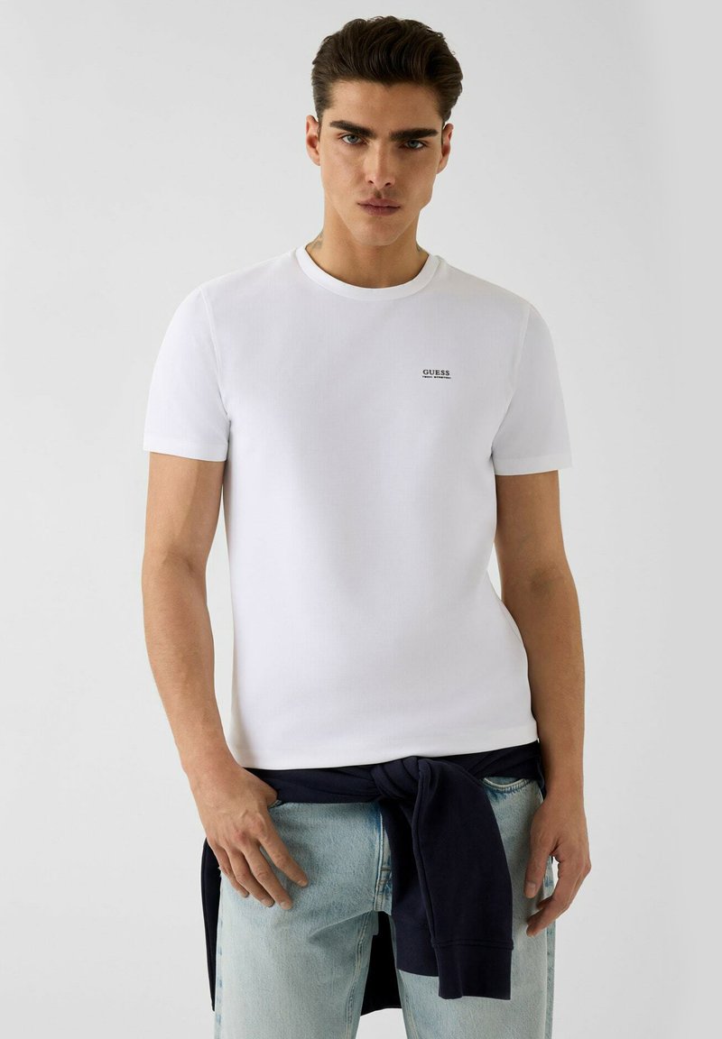 Άνδρας που φοράει λευκό t-shirt Guess και ανοιχτό μπλε τζιν με ένα σκούρο φούτερ δεμένο γύρω από τη μέση, πόζαρε μπροστά σε απλό φόντο.