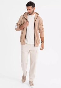 Felpa beige con zip e cappuccio regolabile, abbinata a una t-shirt bianca e pantaloni cargo beige chiaro. Presenta scarpe da ginnastica beige slip-on.