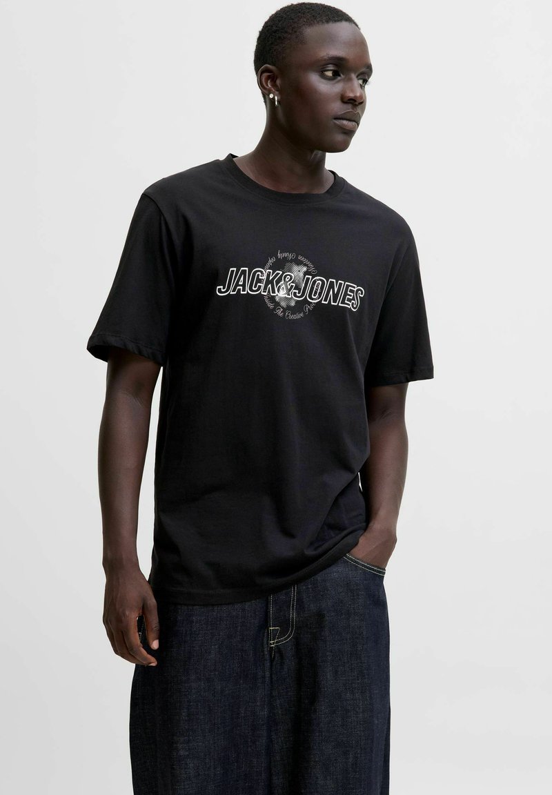 Noor mees, kelles on must "JACK & JONES" t-särk ja tumehallid teksad, seisab ühe käega taskus ja vaatab kõrvale.