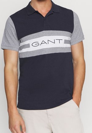 Marineblå polo skjorte med lysegrå ærmer, med et horisontalt stribet middelstykke og "GANT" logo i fed sort tekst. Korte ærmer, klassisk krave.