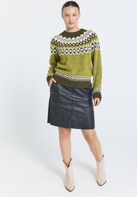 Grön stickad tröja med ett vitt och svart geometriskt mönster, matchad med en svart läderkjol och beiga ankelskor. Slät textur.