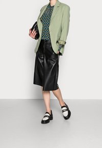 Ljusgröna skräddarsydda kavaj över en mönstrad blåsvart topp, ihop med svarta lädershorts och svart-vita loafers. Släta texturer totalt.