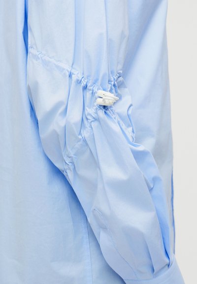 Γαλάζιο πουκάμισο με μακριά μανίκια, με ελαστική σφίγξη και λευκό κούμπωμα με κουμπί που σφίγγει το ύφασμα στο μανίκι κοντά στον αγκώνα.