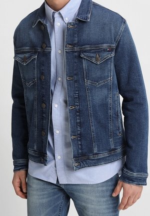 Mand iført en mørkeblå denimjakke over en lyseblå skjorte med knapper og lyseblå jeans, stående med hænderne afslappet langs siden.