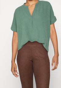 Haut vert à manches courtes avec un col en V, en tissu doux. Associé à des leggings marron côtelés. L'ourlet est légèrement arrondi à la taille.