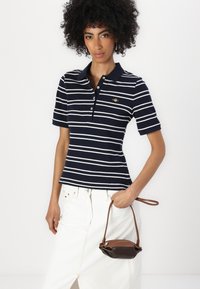 Pánské polo tričko s krátkým rukávem a límečkem v námořnické a bílé pruhované variantě. Model drží malou hnědou a černou kabelku. Tričko je v slim fit střihu a má knoflíkovou légu.
