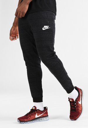 Osoba nosząca czarne spodnie dresowe Nike i czerwone buty sportowe Nike z białymi skarpetkami, stojąca na jednolitym jasnym tle.