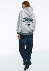 Felpa grigia con cappuccio con scritta sulla schiena, abbinata a pantaloni in denim blu scuro con accenti metallici circolari. Vestibilità comoda, stile casual.