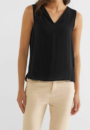 Vrouw draagt een mouwloze zwarte top met V-hals in combinatie met beige broek, staand tegen een effen lichte achtergrond.