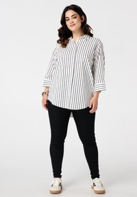 Vrouw in een wit shirt met lange mouwen met zwarte verticale strepen, zwarte broek en witte sneakers, staande tegen een plain witte achtergrond.