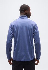 Άνδρας με την πλάτη γυρισμένη, φορώντας μια μπλε αθλητική μπλούζα με μακριά μανίκια και μαύρο παντελόνι, μπροστά σε απλό ανοιχτόχρωμο φόντο.