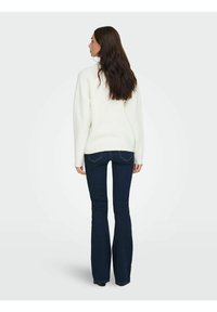 Pull blanc surdimensionné avec poignets et ourlet côtelés, associé à un jean évasé bleu foncé. La texture semble douce et confortable.