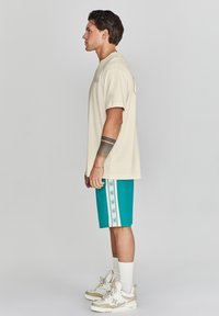 Camiseta beige, pantalones cortos azul verdoso con rayas laterales blancas que presentan logotipos, y zapatillas de color crema. El modelo está de lado, mostrando el ajuste del conjunto.