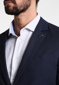 Marinblå blazer i texturerat tyg, med slitsad krage, en framficka och en silveraccentpin på kragen, bärs över en ljus skjorta.