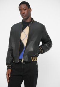 Μαύρο δερμάτινο μπουφάν ταγέρ με ραμμένο γιακά, χρυσές λεπτομέρειες και πλεκτή μέση με τη λέξη "VERS". Φορεμένο πάνω από ένα πουλόβερ με σχέδιο.