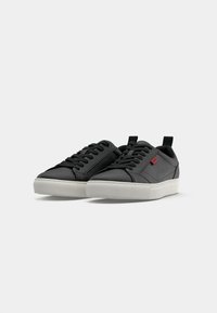 Zwarte low-top sneakers met een gladleren bovenwerk, witte rubberen zool, vetersluiting en een rood logoaccent aan de zijkant.