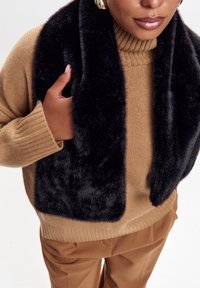 Kvindelig iført tan sweater og bukser, accessorized med guldringe og holder et sort kunstpels tørklæde om halsen.