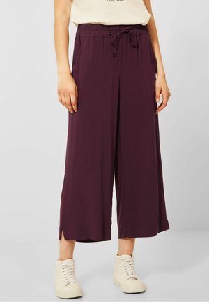 Femme portant un pantalon ample couleur prune à jambes larges avec une taille à cordon et des baskets montantes blanc cassé.