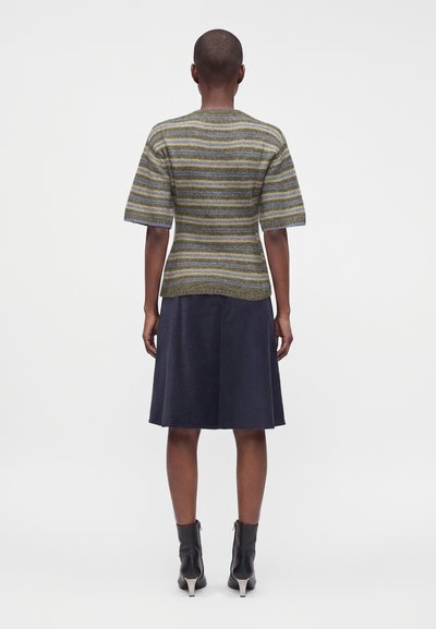 Personne portant un haut tricoté rayé à manches courtes, une jupe foncée mi-longue et des bottines noires, debout de dos sur un fond uni.